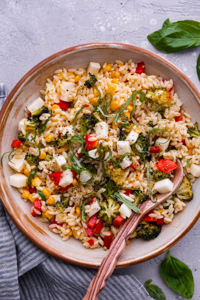This Orzo Pasta Salad is filled with fresh veggies and zesty lemon flavor that will excite your tastebuds. It's perfect for your next potluck, party, or a refreshing and unique meal this summer. You're going to want to save this recipe for later! Orzo pasta salad in a bowl with a wooden spoon.