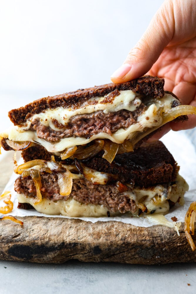 Don't underestimate a homemade patty melt! This is no average sandwich- it's crispy bread filled to the brim with a homemade patty and LOTS of cheese. To top it off, it's got savory and sweet caramelized onions that just make your mouth water. A hand holding a patty melt that's been cut in half.