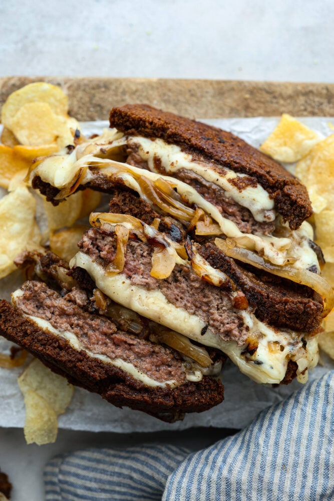 Don't underestimate a homemade patty melt! This is no average sandwich- it's crispy bread filled to the brim with a homemade patty and LOTS of cheese. To top it off, it's got savory and sweet caramelized onions that just make your mouth water. Patty melt sandwiches cut in half with potato chips around them.