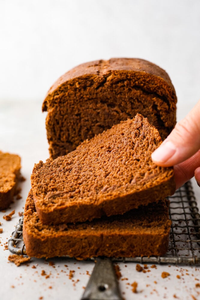 This pumpernickel bread is full of rich and hearty flavor. With three types of flour and brown sugar and molasses, you won't believe how much you can enjoy this nutrient-rich bread! To top it all off, it has cocoa powder to take things to a whole other level. A hand picking up a slice of pumpernickel bread.