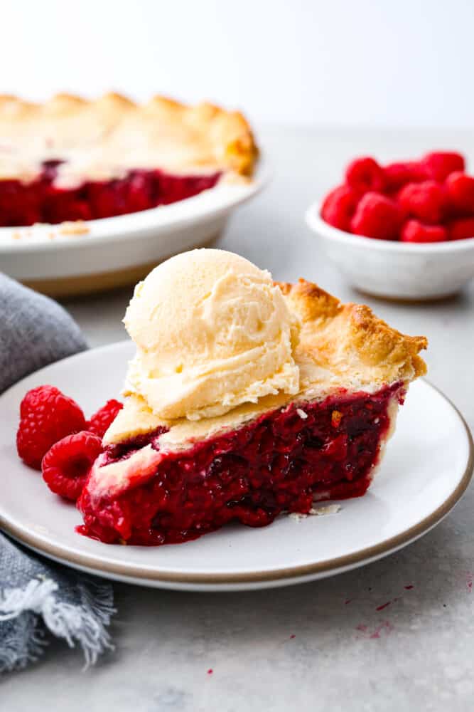 Raspberry pie is a timeless dessert that has been enjoyed for generations, and for good reason. With its flaky, buttery crust and tangy-sweet filling bursting with fresh, juicy raspberries, it’s the perfect balance of flavors and textures. And while store-bought options are convenient, nothing beats making it from scratch! A slice of raspberry pie on a stoneware plate served with a scoop of vanilla ice cream on top.