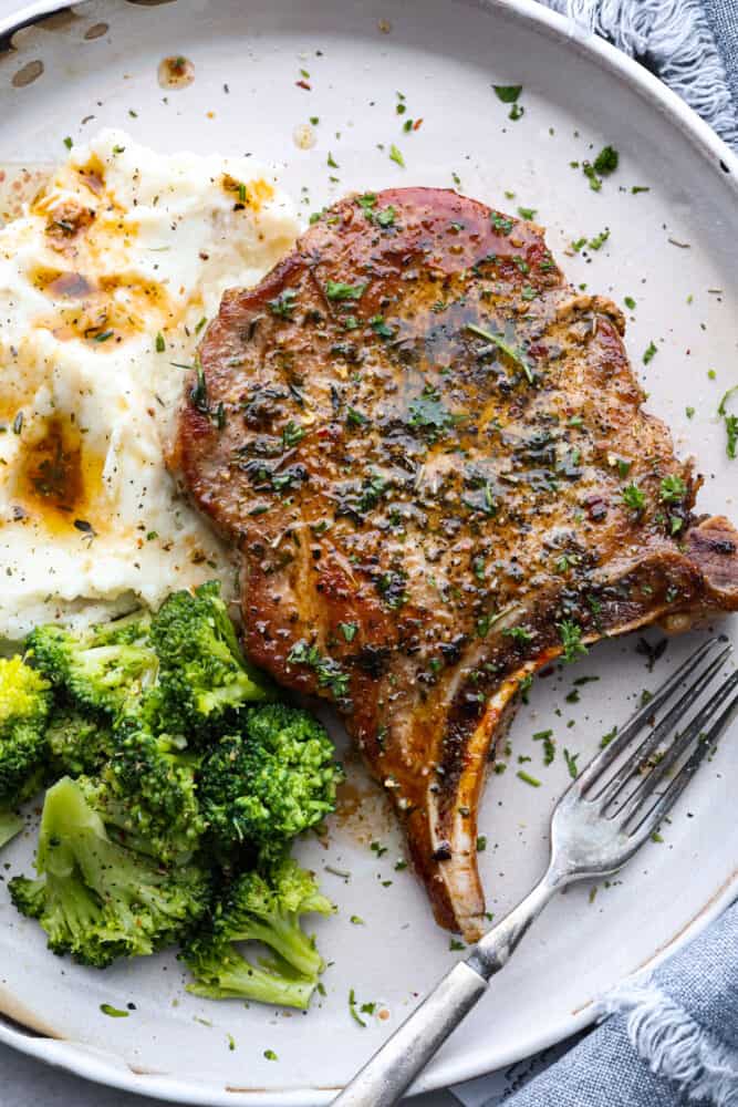 Cast Iron Skillet Pork Chops are a quick and delicious way to serve a satisfying gourmet meal. Bone in pork chops are coated with a garlic herb mixture and seared in a seasoned cast iron skillet for the juiciest and most flavorful pork chop you've ever had! A cast iron skillet pork chop on a plate with mashed potatoes and broccoli.