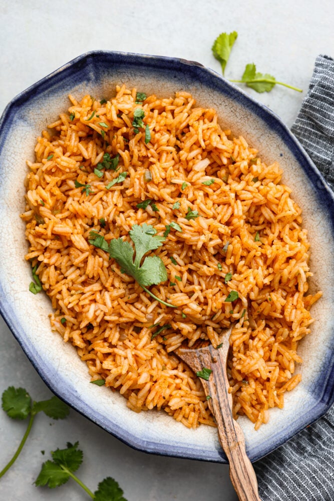 This Spanish rice delivers warm and exciting flavors that complement any meal! This tastes just like your favorite restaurant rice and is delicious served with any protein or Spanish inspired dish. Spanish rice in a serving dish with a wooden spoon and fresh cilantro on top.