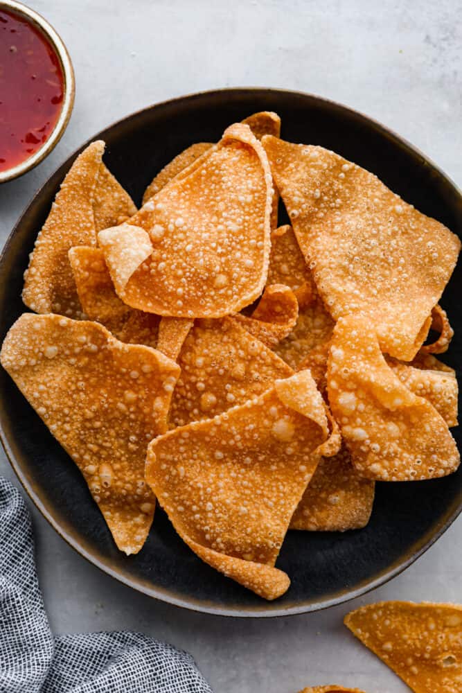 Get ready to up your snack game, because I've got a game-changing recipe for you: wonton chips are where it's at! These golden, crispy triangles are the perfect vessel for all your favorite dips, from guacamole to hummus to sweet and sour sauce. Once you get a taste of that heavenly crunch, you'll wonder how you ever lived without them. Top-down view of wonton chips in a black bowl.