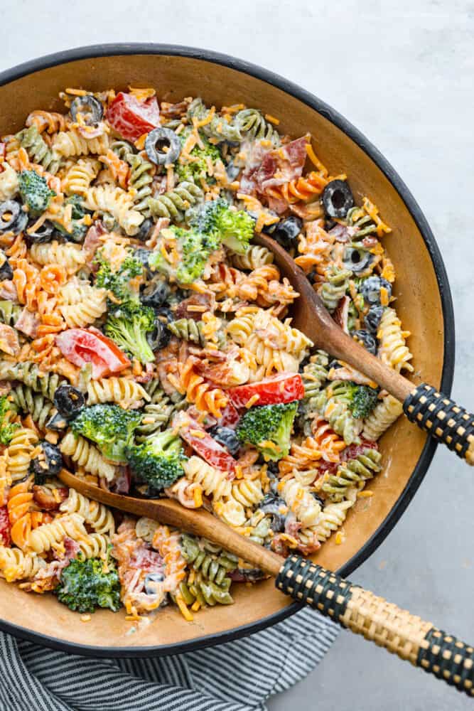 This bacon ranch pasta salad is filled with colorful veggies and perfectly contrasting textures and flavors! Every bite is so satisfying it will be an incredible hit with anyone lucky enough to enjoy it! A bowl of ready to eat bacon ranch pasta sale with 2 wooded spoons in the bowl.