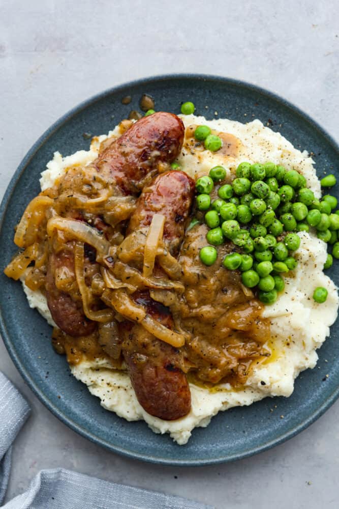 Bangers and mash, a classic British dish, is hearty comfort food at its finest! You'll love these fluffy mashed potatoes and flavorful pork sausages all smothered in a caramelized onion gravy. There's a reason why it's an English favorite! A gray plate topped with mashed potatoes, 2 sausages, onion gravy, and peas.