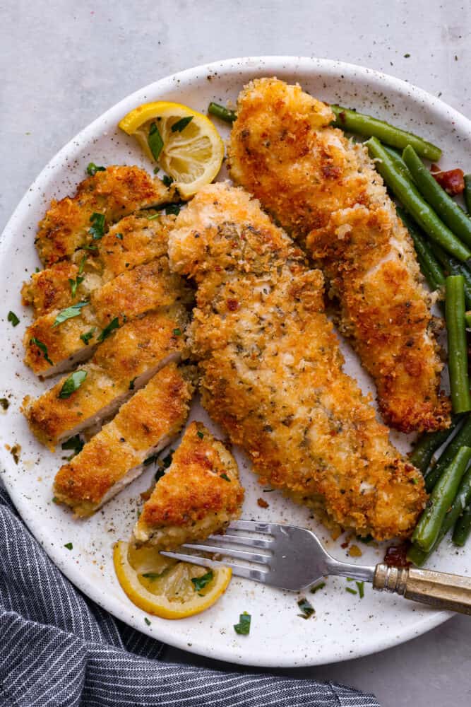 Deliciously seasoned chicken cutlets are breaded and pan-fried until golden brown and crispy. Chicken Milanese is a classic Italian recipe that is fast, easy, and sure to be a weeknight favorite! Top view of chicken Milanese on a white plate. One of the cutlets is sliced with a fork on the plate. Green beans and lemon slices are next to the chicken.