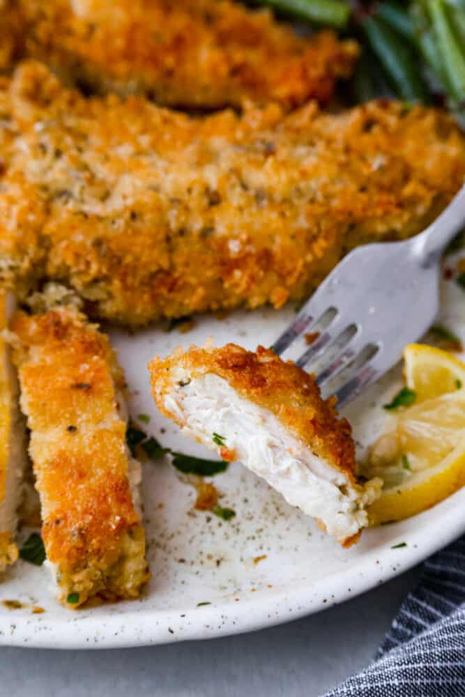 Deliciously seasoned chicken cutlets are breaded and pan-fried until golden brown and crispy. Chicken Milanese is a classic Italian recipe that is fast, easy, and sure to be a weeknight favorite! Close up view of chicken Milanese on a white plate. A piece of chicken is on the fork. Green beans and lemon slices are next to the chicken.