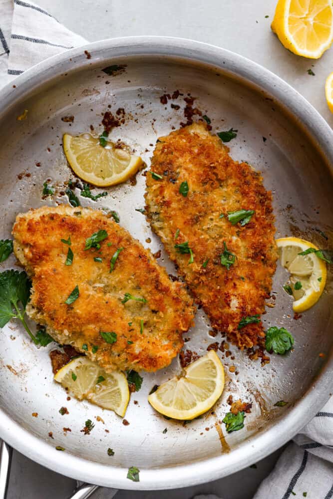 Deliciously seasoned chicken cutlets are breaded and pan-fried until golden brown and crispy. Chicken Milanese is a classic Italian recipe that is fast, easy, and sure to be a weeknight favorite! Top view of chicken Milanese in a silver skillet. Chopped parsley and lemon slices are garnished on top of the chicken.