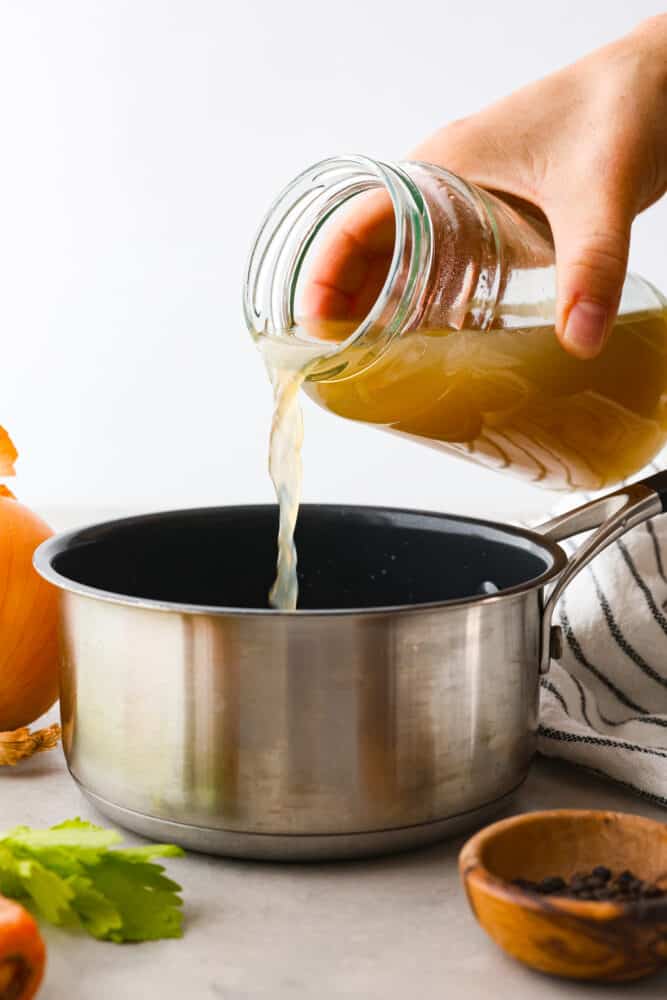 This homemade chicken broth recipe is a keeper and you will love all the many uses you can get out of it! Chicken broth is a staple cooking ingredient used in soups, casseroles, sauces, and many more everyday recipes. Side view of chicken broth being poured into a silver pot. Vegetables and peppercorns are next to the pot.