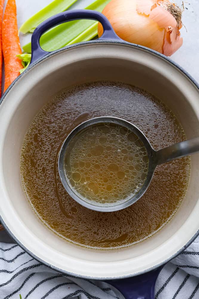 This homemade chicken broth recipe is a keeper and you will love all the many uses you can get out of it! Chicken broth is a staple cooking ingredient used in soups, casseroles, sauces, and many more everyday recipes. Top view of a ladle lifting up chicken broth from a blue pot. Vegetables and a towel are scattered on the counter around the pot.