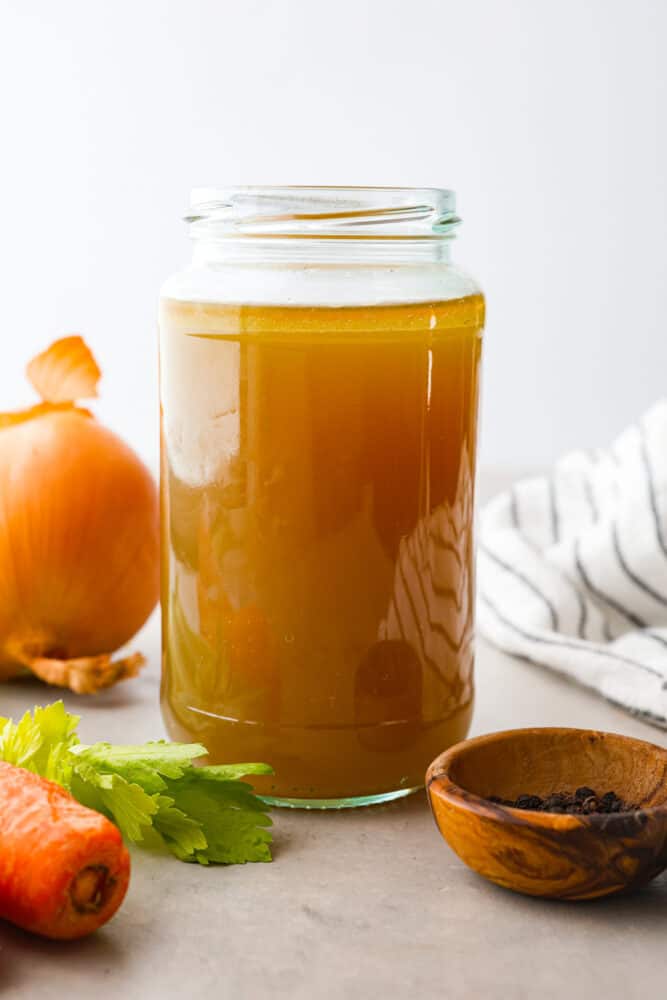 This homemade chicken broth recipe is a keeper and you will love all the many uses you can get out of it! Chicken broth is a staple cooking ingredient used in soups, casseroles, sauces, and many more everyday recipes. Close up view of chicken broth in a large. mason jar with onion, celery, and carrots scattered next to the jar.