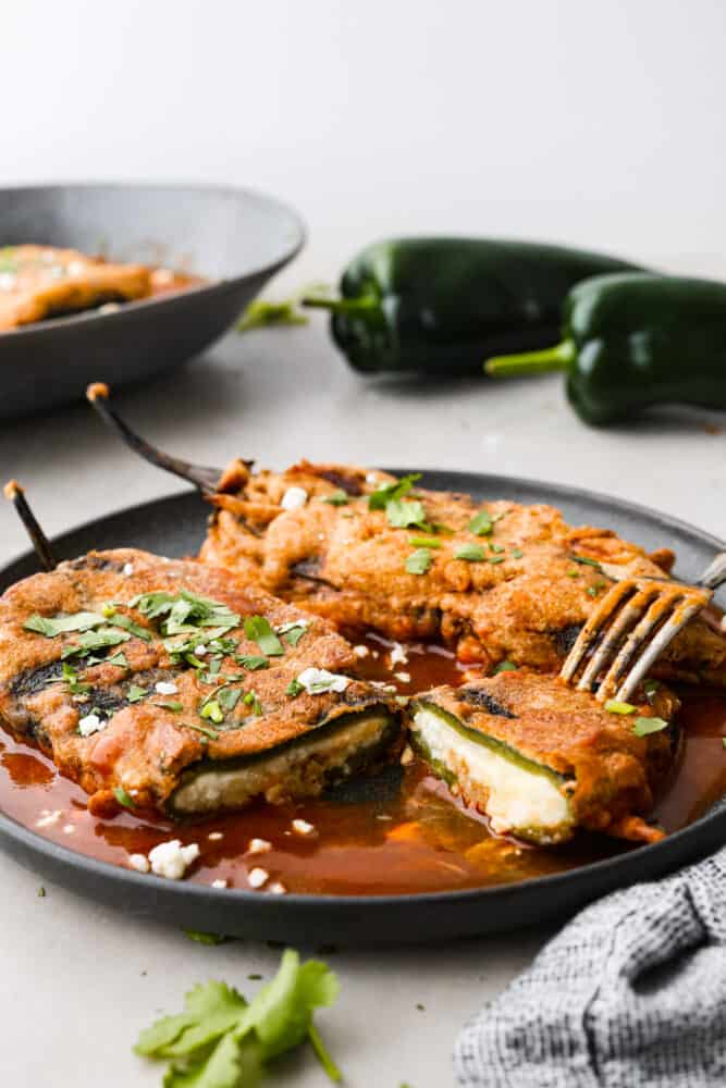 Chile rellenos feature roasted poblano peppers stuffed with a delightful blend of queso fresco and Monterey jack cheese, dipped in a light and fluffy batter, then fried to golden perfection. With its crispy exterior and gooey, cheesy interior, this Mexican appetizer will leave you craving for more. 2 chile rellenos on a plate, with one cut in half.