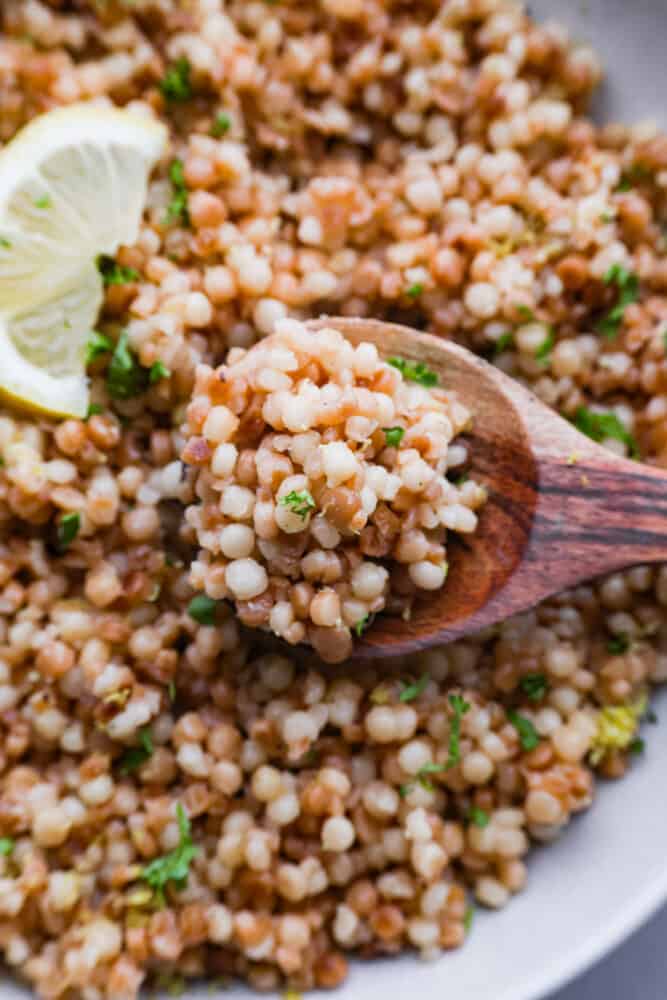 Looking to add a burst of Mediterranean flavor to your dining table? This delicious Israeli couscous is the answer! With its aromatic blend of olive oil, lemon zest, garlic, and fresh parsley, you won't be able to get enough. It's the perfect side dish for summer! Closeup of a wooden spoon.