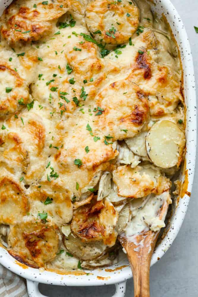 Scalloped potatoes are thinly sliced layered potatoes baked in a creamy garlic and herb sauce, making them tender and flavorful. Hearty and easy to make, this is the perfect side dish to any meal! Hero image of a scoop of scalloped potatoes being taken out of a white baking dish.