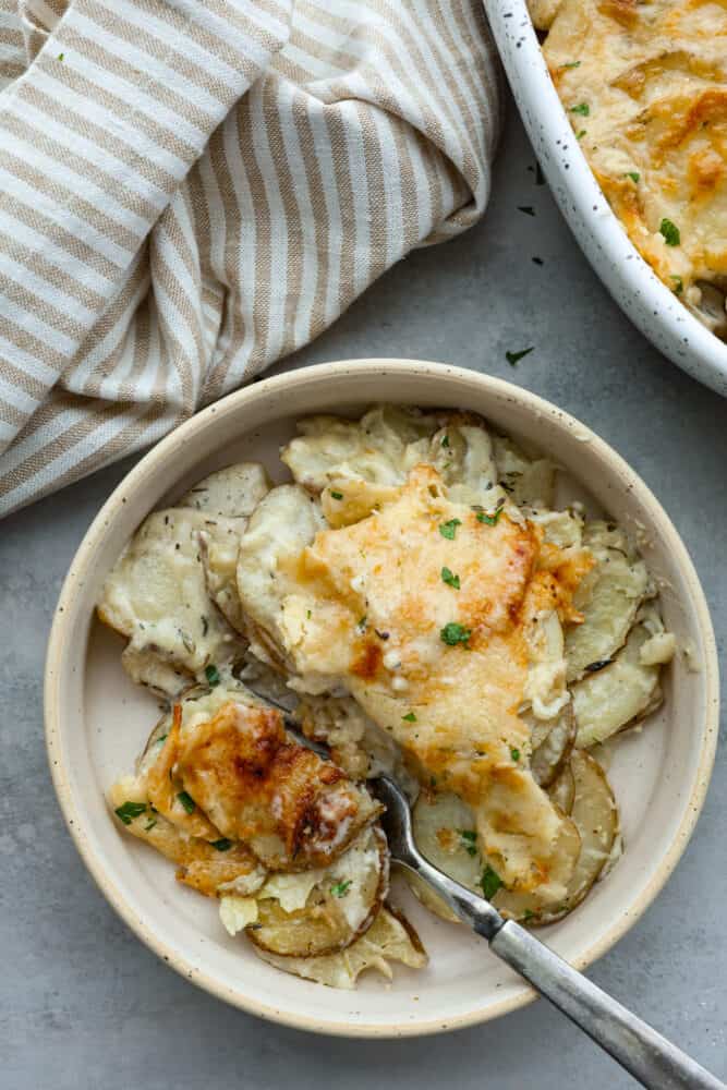 Scalloped potatoes are thinly sliced layered potatoes baked in a creamy garlic and herb sauce, making them tender and flavorful. Hearty and easy to make, this is the perfect side dish to any meal! A serving of potatoes in a stoneware bowl.