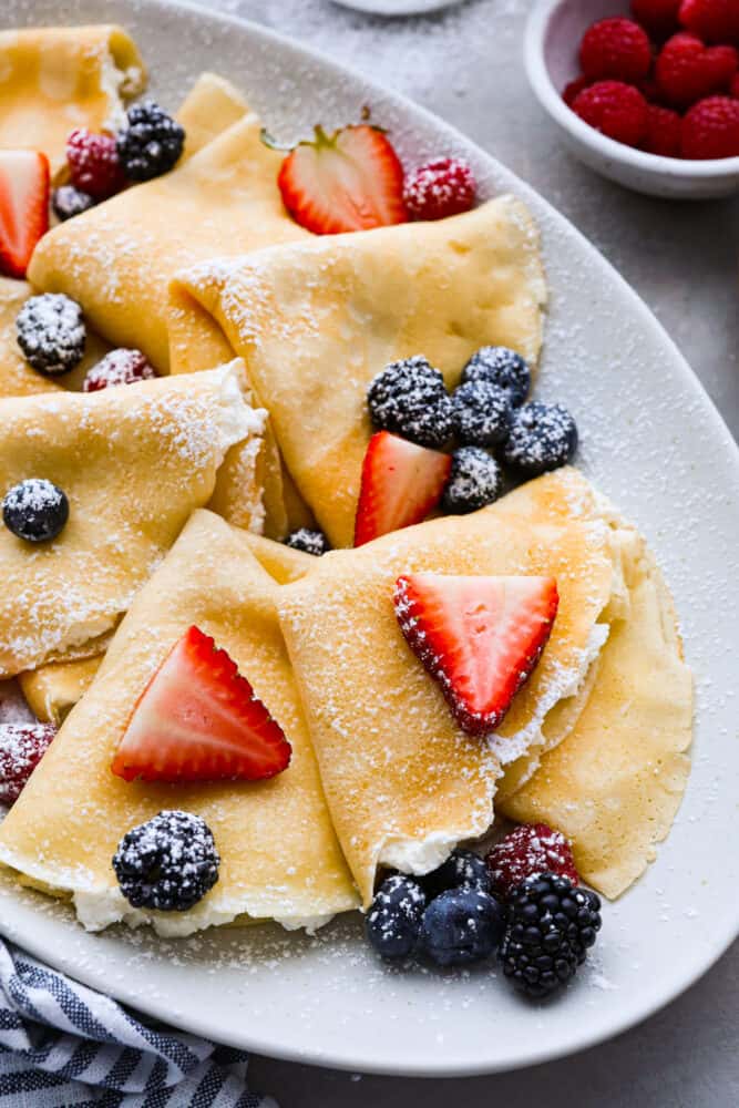 Perfect Crepes are a light and thin pancakes that is simply blended batter that makes a sweet or savory breakfast for any day. The toppings are endless! Close up of folding crepes on a gray platter. The crepes are dusted with powdered sugar and topped with berries.
