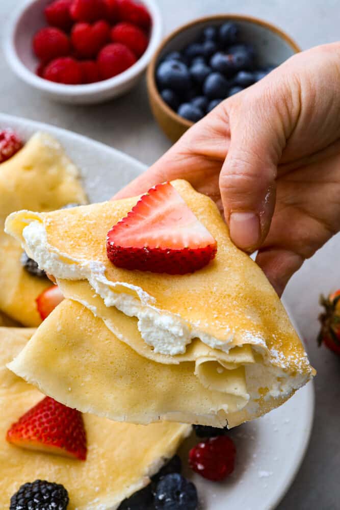 Crepes are thin and delicate French pancakes served with endless topping options such as fruit, sauces, and whipped cream. The batter is easily blended and cooked into tender foldable perfect crepes. A folded crepe being held with filling on the inside and berries on top.