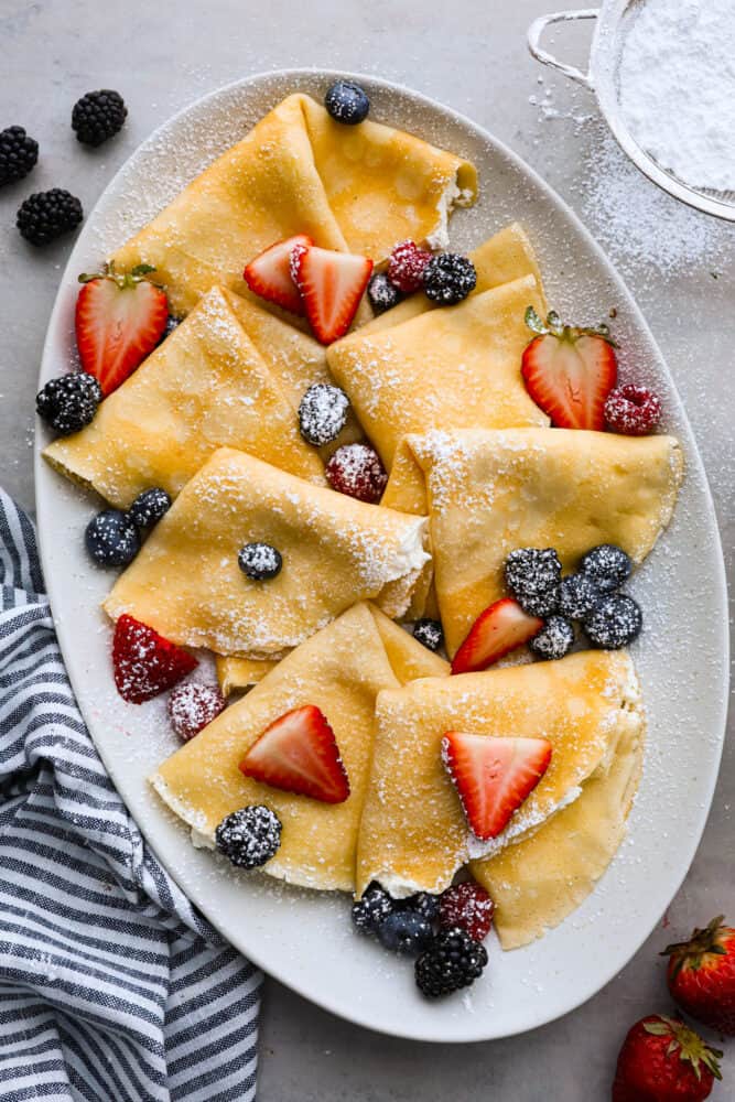 Perfect Crepes are a light and thin pancakes that is simply blended batter that makes a sweet or savory breakfast for any day. The toppings are endless! Overhead view of folded crepes on a gray platter dusted with powdered sugar and topped with fresh berries.