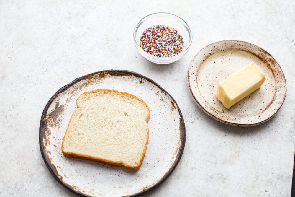 Fairy bread is the easiest treat to make and the best way to add a little magic to your day! These slices of buttery, sprinkled goodness are a classic Australian recipe, and sure to win your kids over. A plate of bread, a bowl of sprinkles, and a stick of butter.