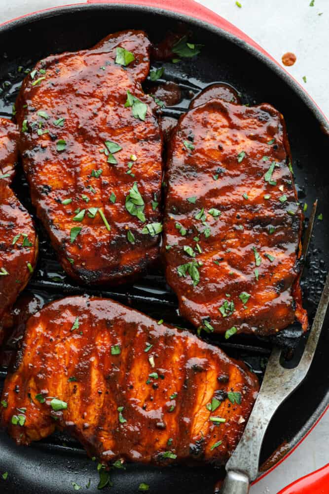 Get ready to take your pork chops to a whole new level with these BBQ Pork Chops. Grilled to perfection, they are juicy, tender, and bursting with tangy goodness. Prepare to impress your taste buds with this simple yet delicious dish. A close up of the post chops in a pan garnished with cilantro.