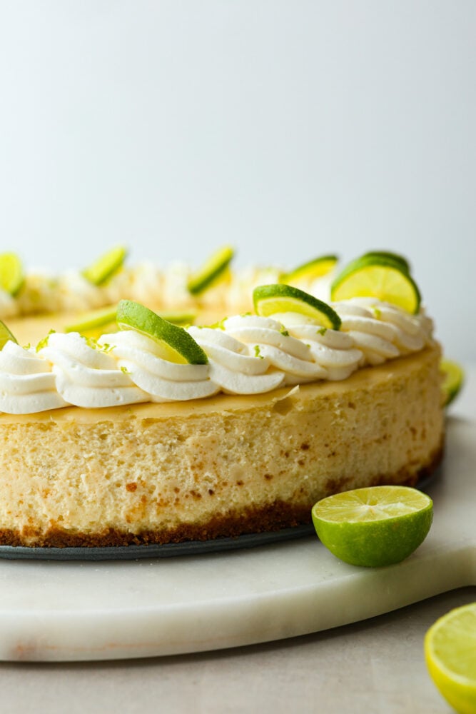 Experience the perfect summer treat with this key lime cheesecake. Made with a scrumptious graham cracker crust and topped with lime zest whipped cream, this creamy but juicy dessert is a refreshing delight! Each bite is the perfect combination of tart and sweet. A whole cheesecake, garnished with whipped cream and sliced limes.