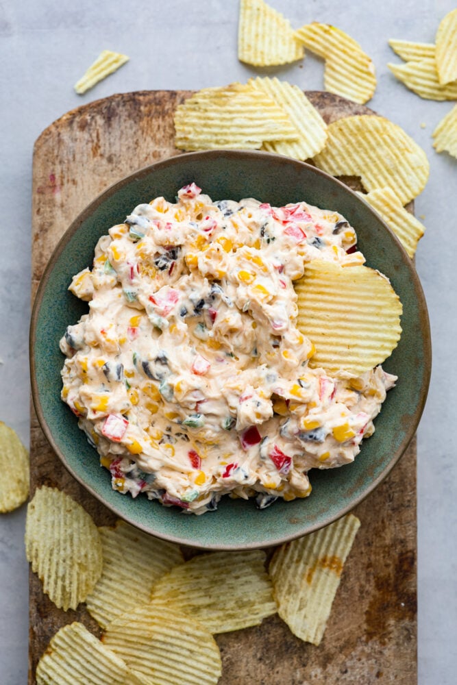 Loaded Creamy Ranch Dip is packed with so many delicious flavors that friends and family won't be able to get enough of it. Perfect for the pool or potlucks! A bowl of creamy ranch dip on a wooden cutting bread with potato chips scattered around.