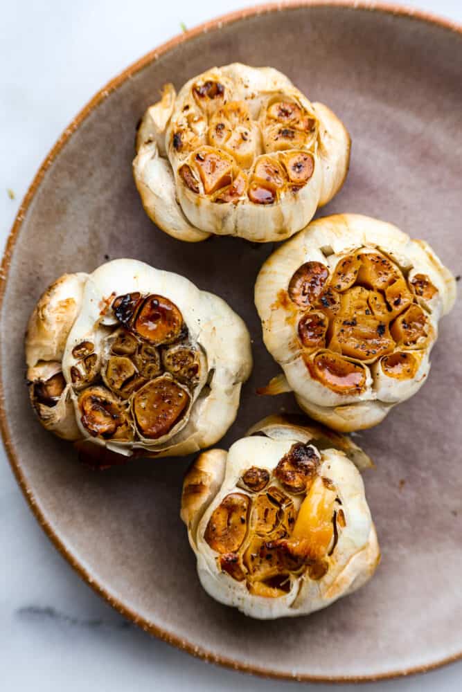If you want to infuse your favorite recipes with savory, nutty flavor, roasted garlic is the way to go! You can easily make some at home in a muffin tin with a drizzle of olive oil on top to enhance the overall flavor! The end result is caramelized, garlic heaven. 4 heads of roasted garlic in a gray dish.
