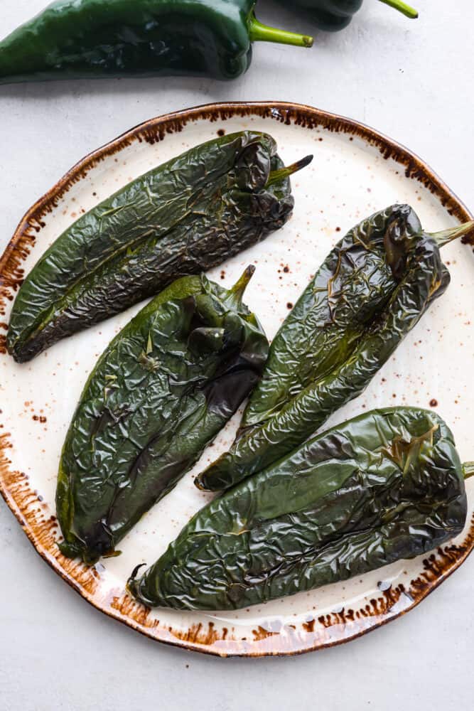 If you're a fan of peppers and haven't tried roasted poblano peppers yet, you're missing out! These peppers have a unique flavor that is smoky, slightly sweet, and just a little bit spicy. Roasting them brings out their full potential, making them perfect for giving your favorite Mexican dishes a little kick. 4 roasted poblano peppers on a white plate.