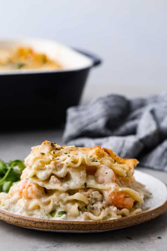 If youโre a seafood lover, you'll be obsessed with this Seafood Lasagna. Iโm talking crab, scallops, and shrimp in creamy, cheesy layers, baked to bubbly perfection. Itโs my go-to dinner when Iโm cooking for seafood fans! Close up view of seafood lasagna on a white plate with the pan of lasagna is in the background.
