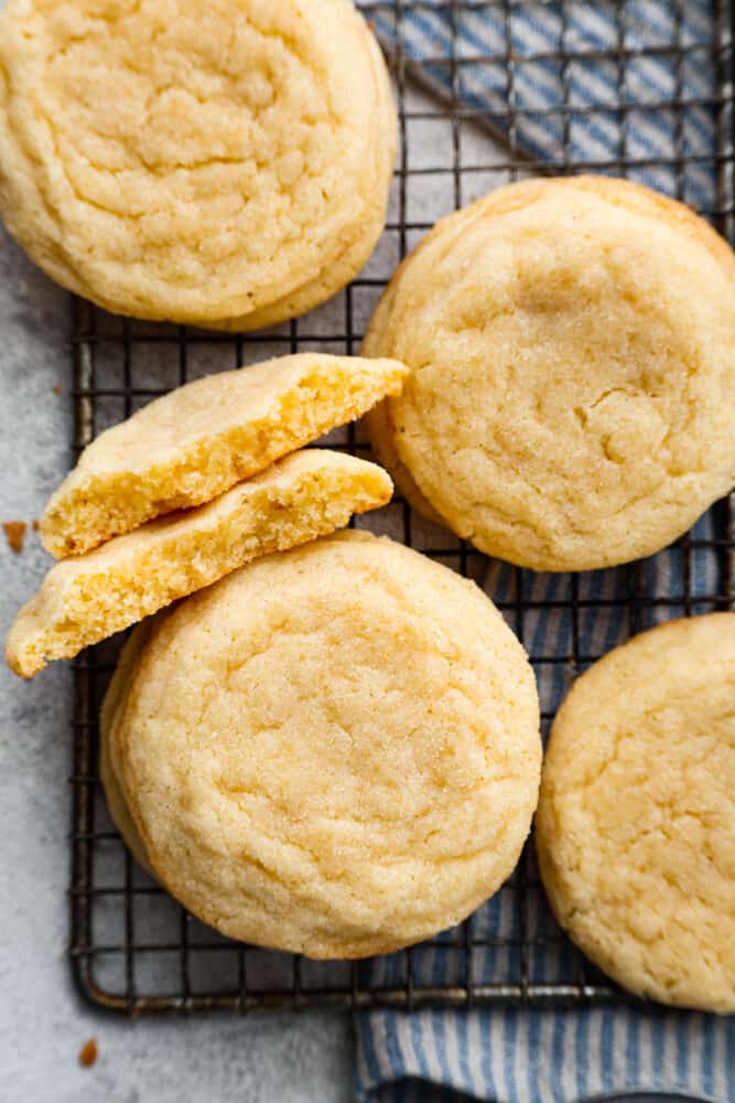 These soft sugar cookies are perfectly chewy on the inside with sweet golden edges on the outside! The drop-style method makes these the easiest sugar cookies you will ever make, and everyone will fall in love with the chewy texture. Close up view of stacked sugar cookies on a cooling rack. Cookies are broken in half and stacked next to each other.