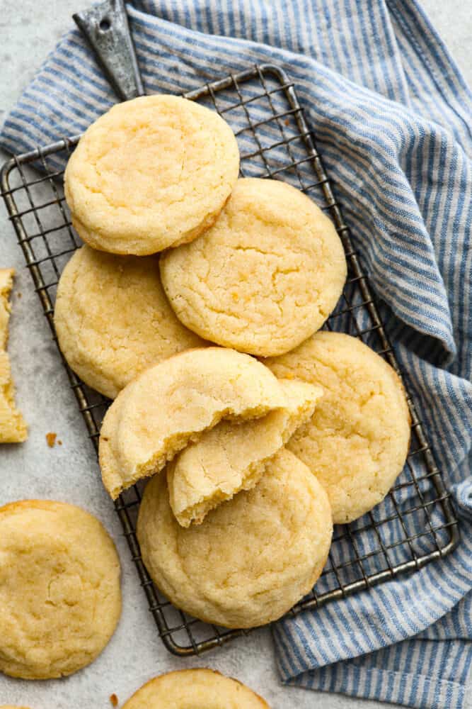 These soft sugar cookies are perfectly chewy on the inside with sweet golden edges on the outside! The drop-style method makes these the easiest sugar cookies you will ever make, and everyone will fall in love with the chewy texture. Close up side view of soft sugar cookies stacked on a cooling rack. The cookie on top of the stack is broken in half with the other half on the counter.