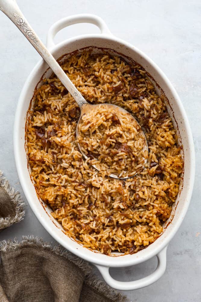 Stick of Butter Baked Rice is soft and fluffy rice that is loaded with sweet, rich flavors from onion soup and butter.ย It is quick and easy to prep and bakes in the oven to perfection! The top view of baked rice in a casserole dish with a large spoon.