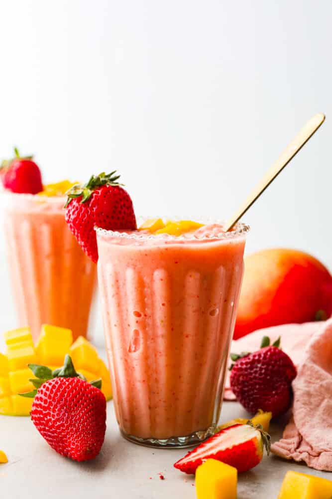 This delightful Strawberry Mango Smoothie recipe is a burst of tropical flavors that will transport you to paradise. With just a handful of ingredients and a blender, you'll be sipping on this refreshing treat in no time. A strawberry mango smoothie in a clear glass with a gold spoon. Strawberries and mangoes are around the cup.