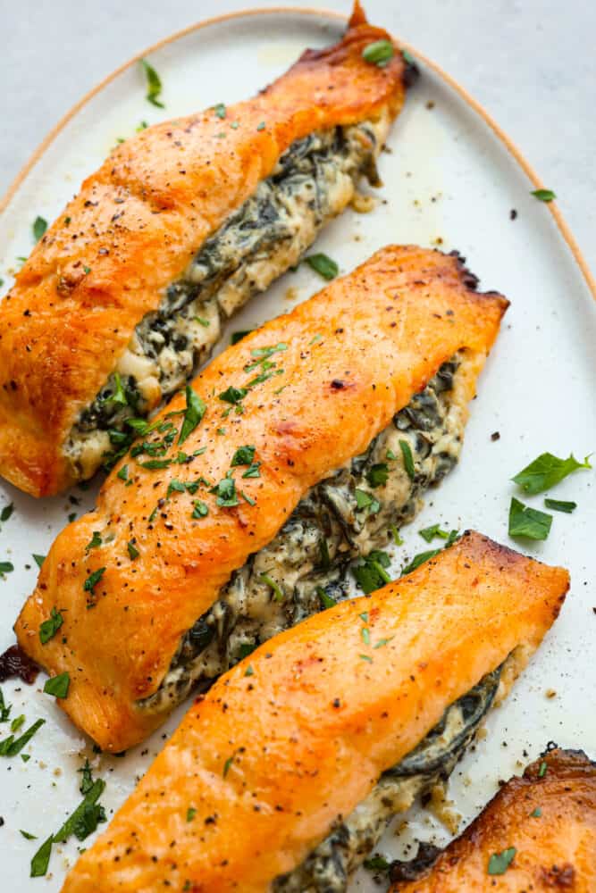 This spinach stuffed salmon is a flavor explosion, with the creamy richness of the cheese and the earthy goodness of the spinach perfectly complementing the tender, flaky salmon. It's a restaurant-quality meal that's ready in just 45 minutes! 3 stuffed salmon filets on a white plate.