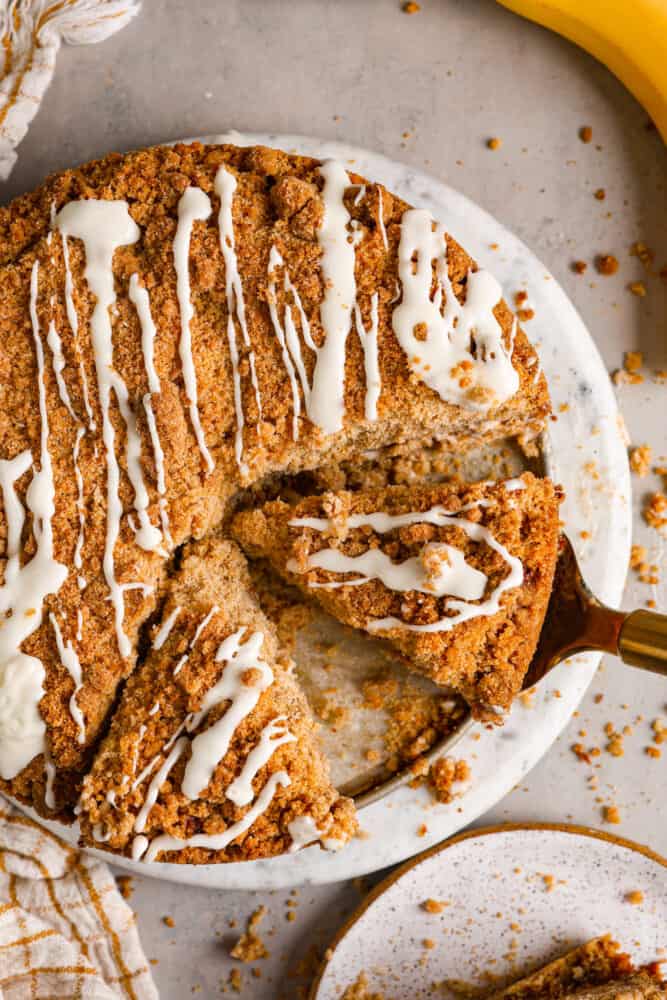 Banana coffee cake is a great way to use up any overripe bananas you might have lying around! With a moist, fluffy, vanilla banana-flavored interior and a buttery streusel topping, this is a unique twist on coffee cake that will have you hooked after the first bite! Top-down view of banana coffee cake, with a slice being served with a cake spatula.