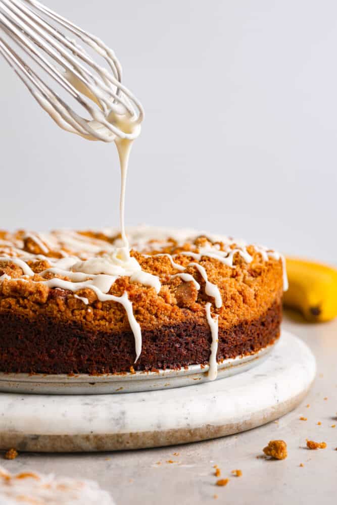 Banana coffee cake is a great way to use up any overripe bananas you might have lying around! With a moist, fluffy, vanilla banana-flavored interior and a buttery streusel topping, this is a unique twist on coffee cake that will have you hooked after the first bite! Glaze being drizzled onto the cake.