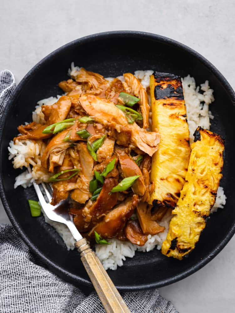 Crockpot huli huli chicken is a dinner you can set and forget, then come back to and enjoy in all of its tropical goodness! Each bite of fork-tender chicken is flavored with the best sauce made of ginger, garlic, ketchup, brown sugar, and pineapple juice. It's the perfect mix of sweet, tangy, and savory! Huli huli chicken served in a black bowl along with some white rice and grilled pineapple.
