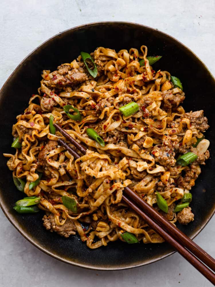 Dan Dan Noodles is a classic Chinese dish made with tender egg noodles and flavorful minced pork tossed in a savory, slightly spicy sauce. It's the perfect comfort food and will have everyone in your family wanting seconds. Noodles served in a black bowl. Chopsticks are shown in the picture.