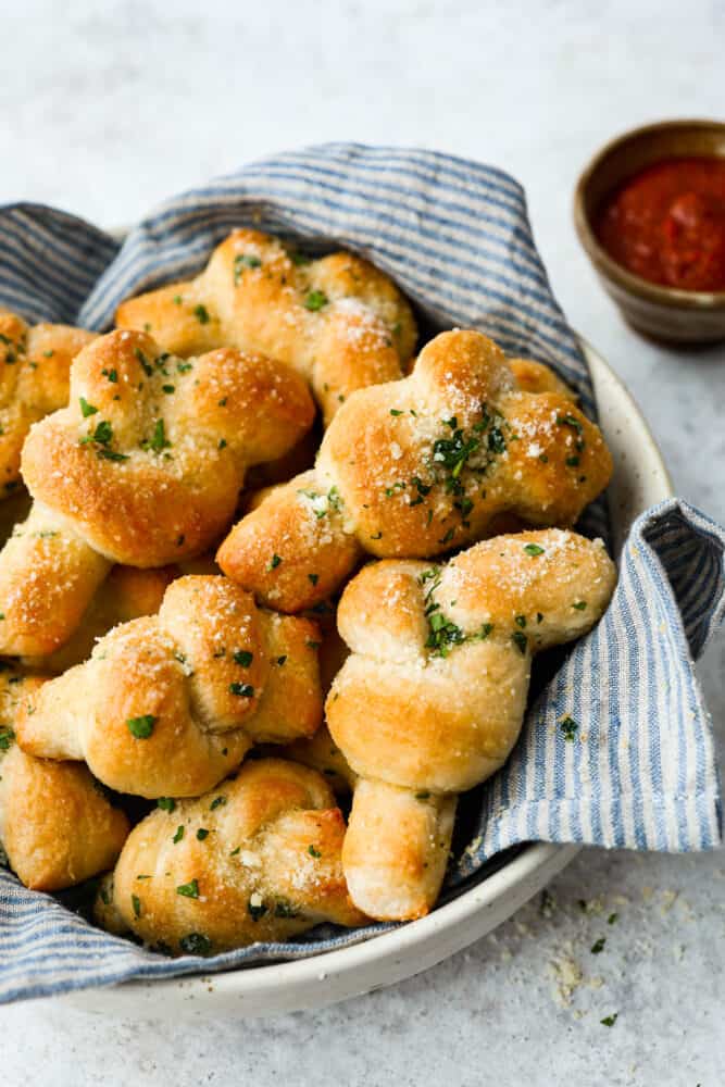 Homemade garlic knots work for any meal and are super easy to make. They're coated with a mouthwatering garlic spread made from butter, garlic powder, parmesan cheese, and fresh herbs! Serve them up with your favorite Italian dish and marinara sauce for dipping. It's the best combination! Hero image of homemade garlic knots in a white serving bowl.
