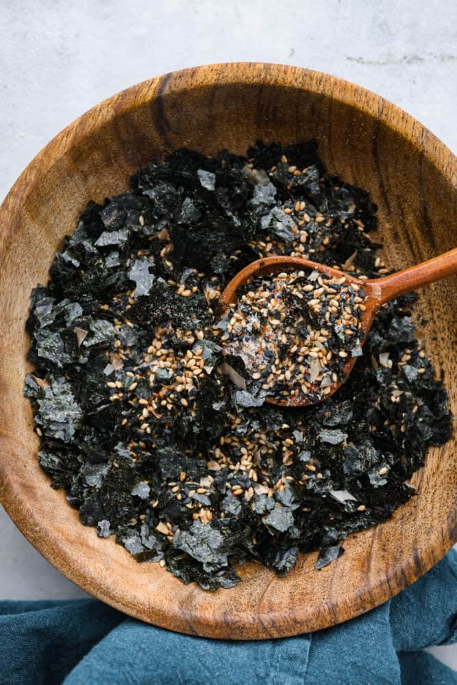 Furikake seasoning is a delightful mix of savory, umami goodness with a hint of saltiness. It's made with a blend of nutty seeds, spices, dried fish, and crispy seaweed- don't knock it 'til you try it! Sprinkle this Japanese condiment over rice, noodles, veggies, or even chips or popcorn to instantly elevate their flavors and add a satisfying crunch. Furikake seasoning in a wooden bowl.