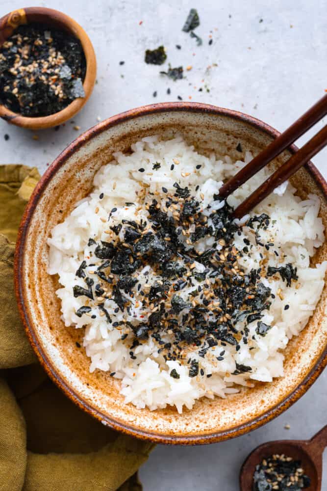 Furikake seasoning is a delightful mix of savory, umami goodness with a hint of saltiness. It's made with a blend of nutty seeds, spices, dried fish, and crispy seaweed- don't knock it 'til you try it! Sprinkle this Japanese condiment over rice, noodles, veggies, or even chips or popcorn to instantly elevate their flavors and add a satisfying crunch. A bowl of rice topped with furikake.