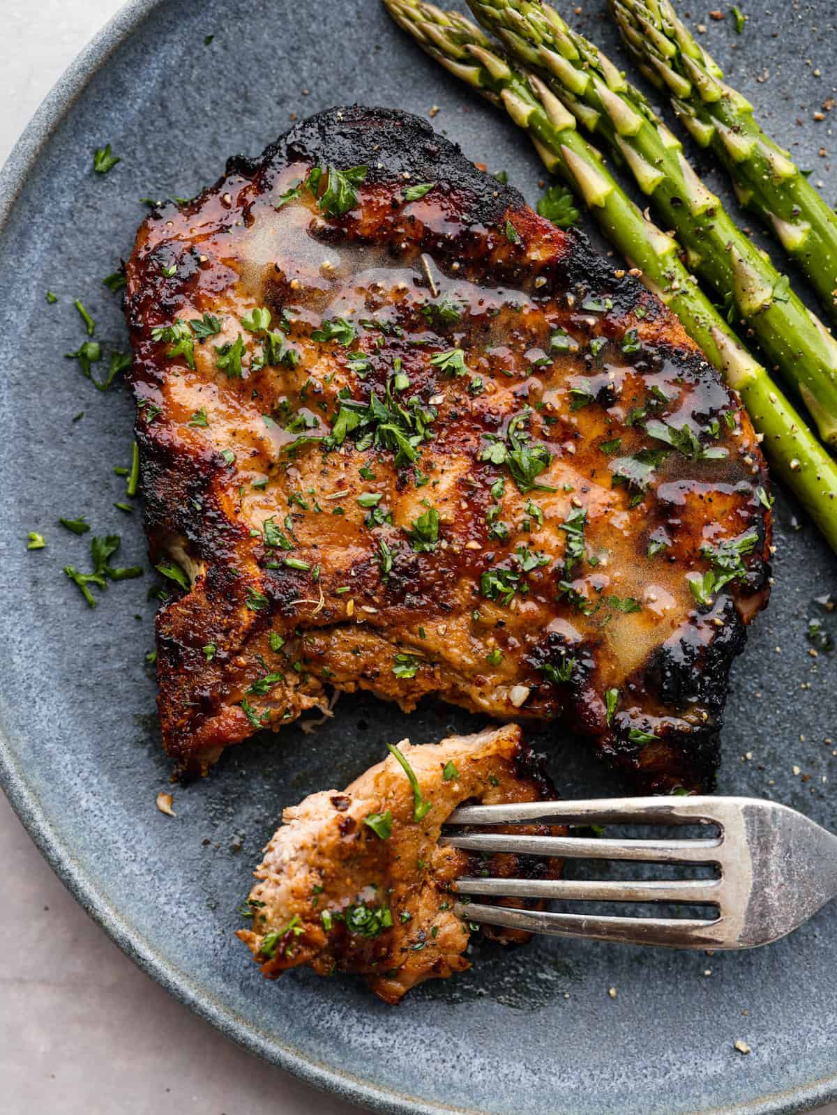 Grilling your pork chops is the best way to make them! These grilled pork chops are marinated until they’re extra flavorful and tender, then cooked to smoky perfection. They’re sure to fly off the table at your next barbecue! A pork chop served with asparagus. There is a bite taken out of it.