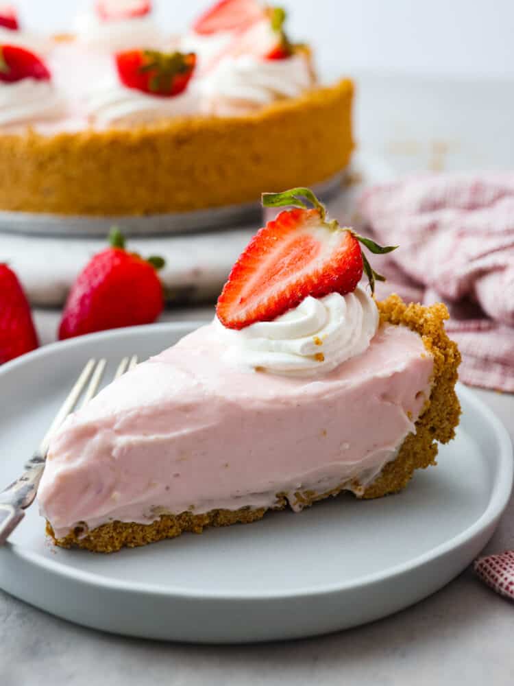 This easy and refreshing no-bake strawberry cheesecake is the perfect summer dessert! The creamy cheesecake filling is mixed with a fresh strawberry puree and nestled inside a sweet graham cracker crust. A close up view of a slice of no-bake strawberry cheesecake on a light blue colored plate.The pie is garnished with whipped cream and a strawberry slice. A fork is on the plate next to the slice.