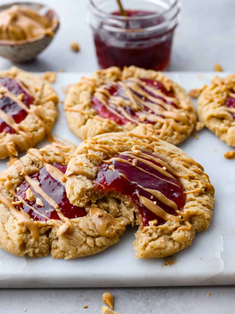 A classic flavor pairing, but in cookie form! These peanut butter and jelly cookies are extra soft and delicious, each with a strawberry jam core and peanut butter drizzle on top. Closeup of a cookie with a bite taken out of it.