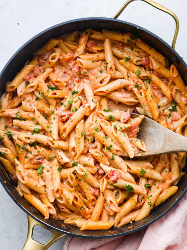 This ultra-delicious Pink Sauce Pasta recipe features penne pasta cooked al dente and tossed in a flavorful pink sauce. The dish is garnished with fresh basil and grated parmesan cheese for an extra touch of deliciousness. A photo of pink sauce pasta in a pan with a wooden spoon.