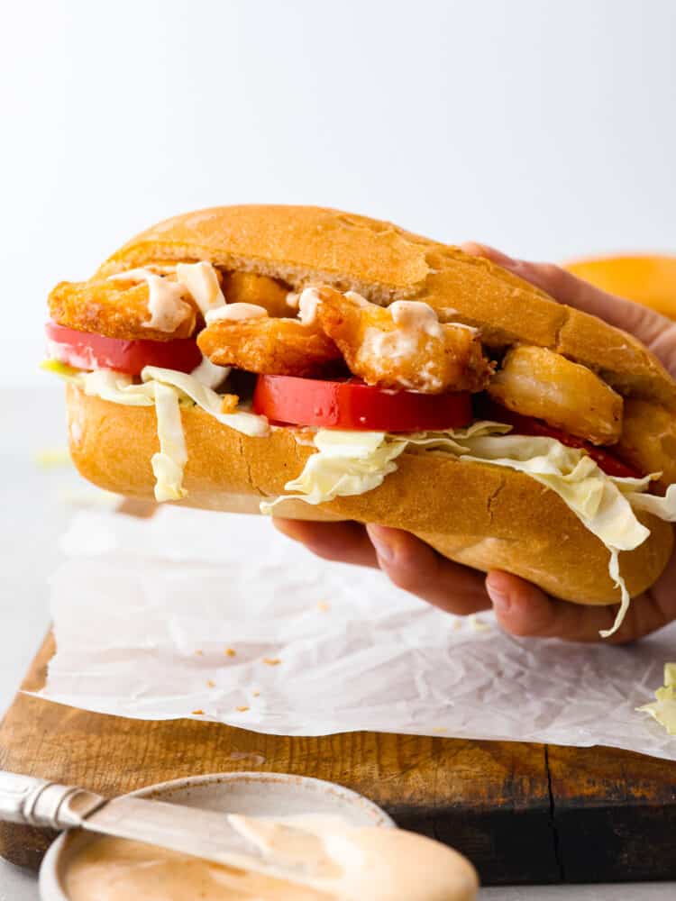 New Orlean's introduced us to the shrimp po boy sandwich and it's loaded with crispy cajun fried shrimp. This southern classic is complete with soft French bread, tomatoes, cabbage, and smothered in a zesty sauce. Close up side view of a hand holding a shrimp po boy sandwich.