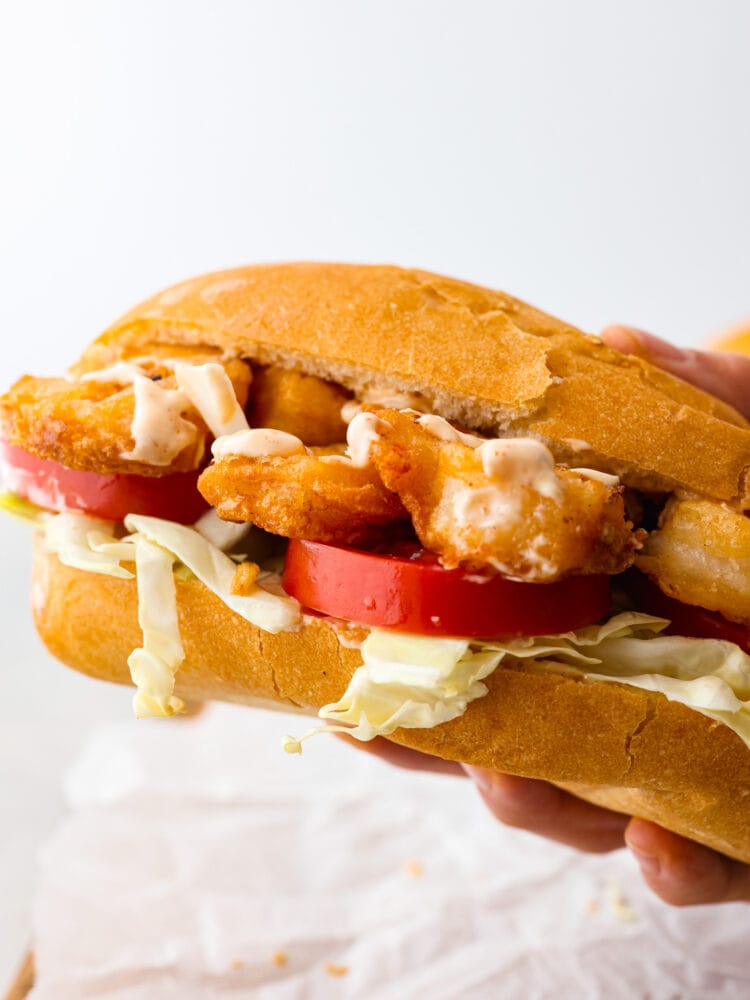 New Orlean's introduced us to the shrimp po boy sandwich and it's loaded with crispy cajun fried shrimp. This southern classic is complete with soft French bread, tomatoes, cabbage, and smothered in a zesty sauce. Close up view of a shrimp po boy sandwich being held up. Crispy fried shrimp, tomatoes, and cabbage are nestled in French bread.