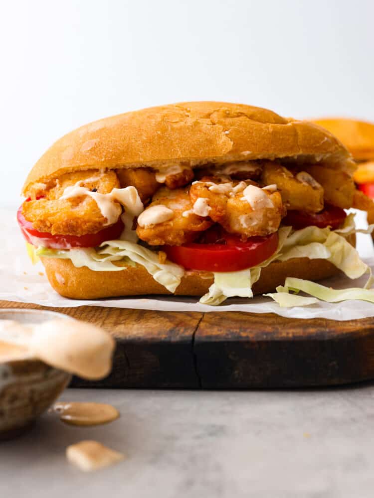 New Orlean's introduced us to the shrimp po boy sandwich and it's loaded with crispy cajun fried shrimp. This southern classic is complete with soft French bread, tomatoes, cabbage, and smothered in a zesty sauce. Close up view of shrimp po boy on a wood board. Cajun aioli is in a bowl next to the sandwich with the sauce dripping off the knife.