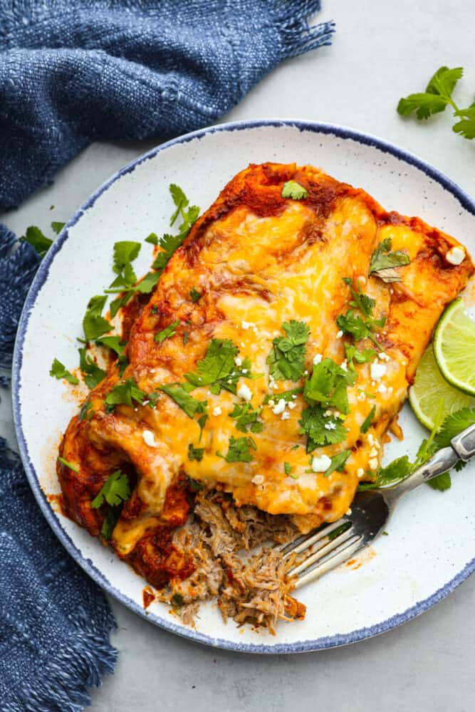 These delicious enchiladas are filled with juicy pork carnitas and smothered in melted cheese. Topped off with homemade red enchilada sauce, this is a dinner that the whole family will go crazy over. Top-down view of pork enchiladas, garnished with cilantro, on a blue and white plate.